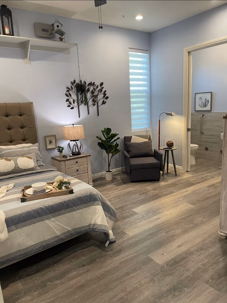 Well-decorated bedroom with a bed, nightstand and lamp, armchair and plants, and an open doorway to a bathroom.