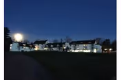 A large white building with multiple sections and windows illuminated at night under a dark blue sky, with a grassy area in the foreground.