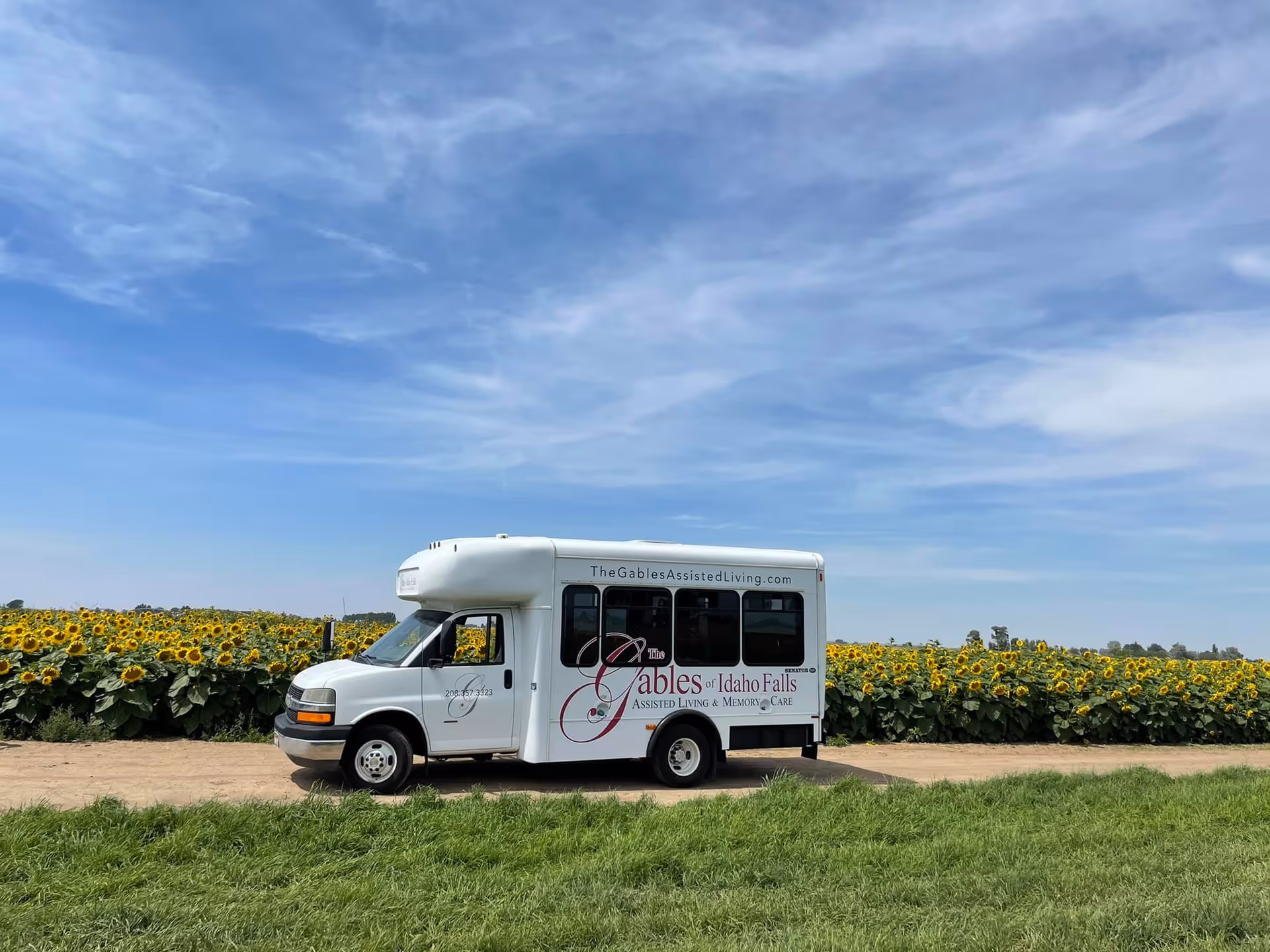 A white shuttle bus from The Gables Assisted Living of Idaho Falls parked on a dirt path next to a large field of blooming sunflowers under a partly cloudy blue sky.