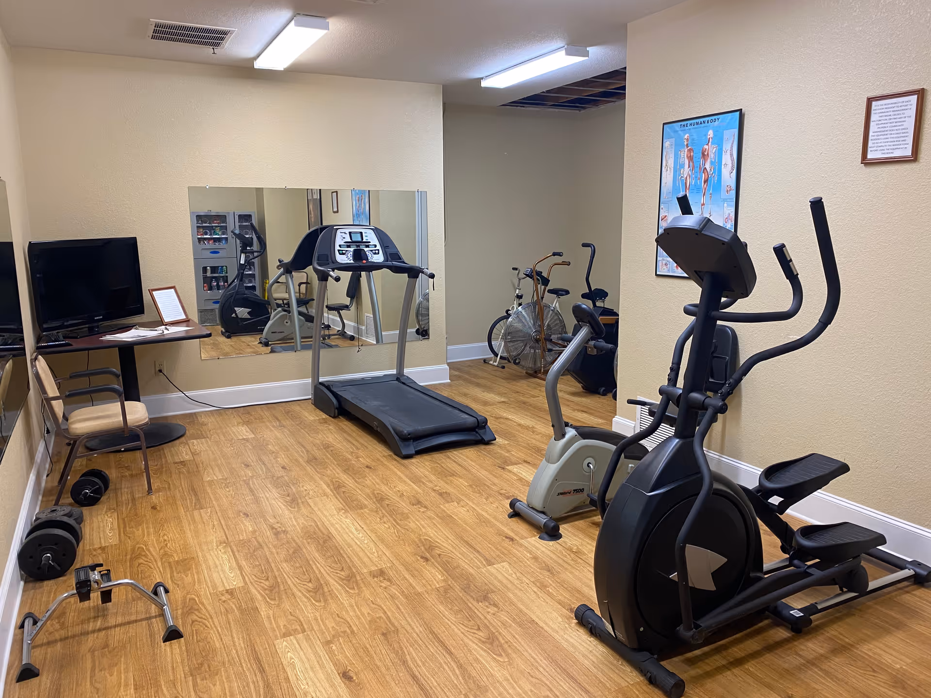 A small fitness room with wooden flooring containing exercise equipment including a treadmill, elliptical machine, stationary bikes, and dumbbells. There is a large mirror on one wall, a TV mounted on another wall with a small table and chair beneath it, and a poster of the human body on the far wall.