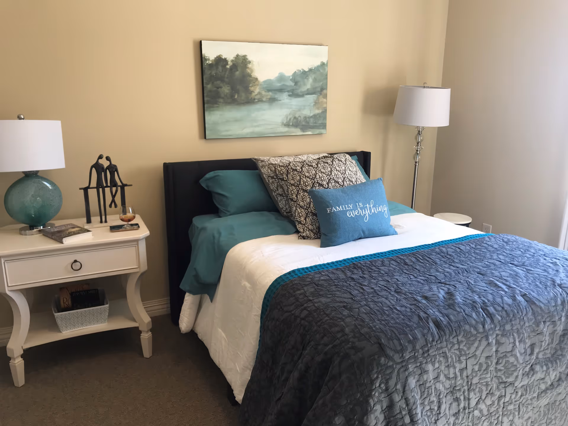 A neatly made bed with teal and white bedding, decorative pillows including one with the text 'FAMILY IS everything', a white nightstand with a green lamp, books, and a small sculpture, and a floor lamp next to the bed in a softly lit bedroom with beige walls and a landscape painting above the bed.