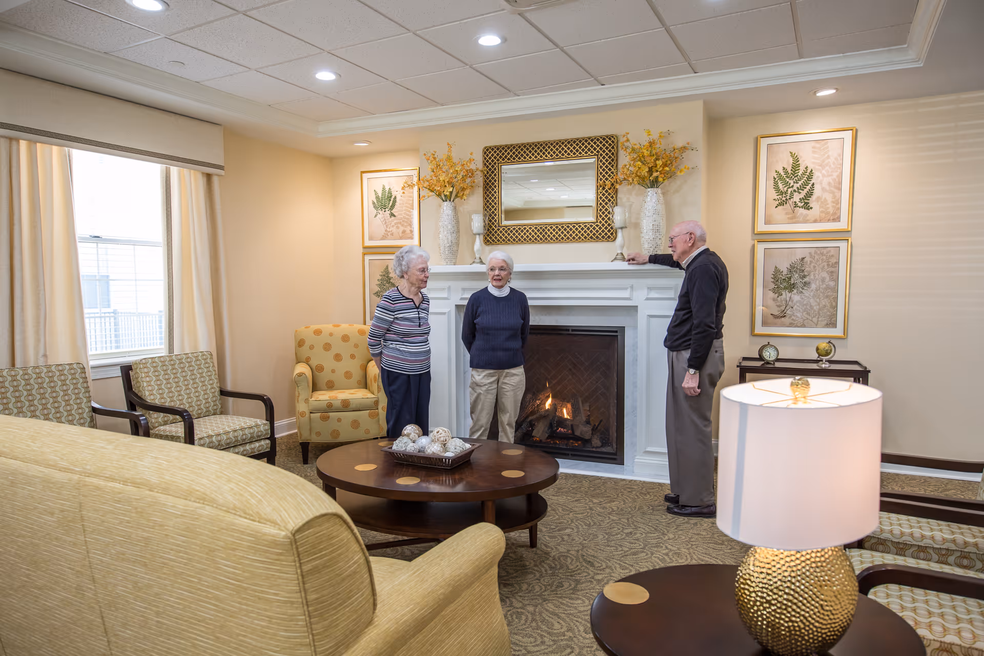 Three elderly people standing and conversing in a cozy living room with a lit fireplace, beige walls, framed botanical prints, and comfortable seating including armchairs and a sofa. A table lamp and decorative items are also visible.