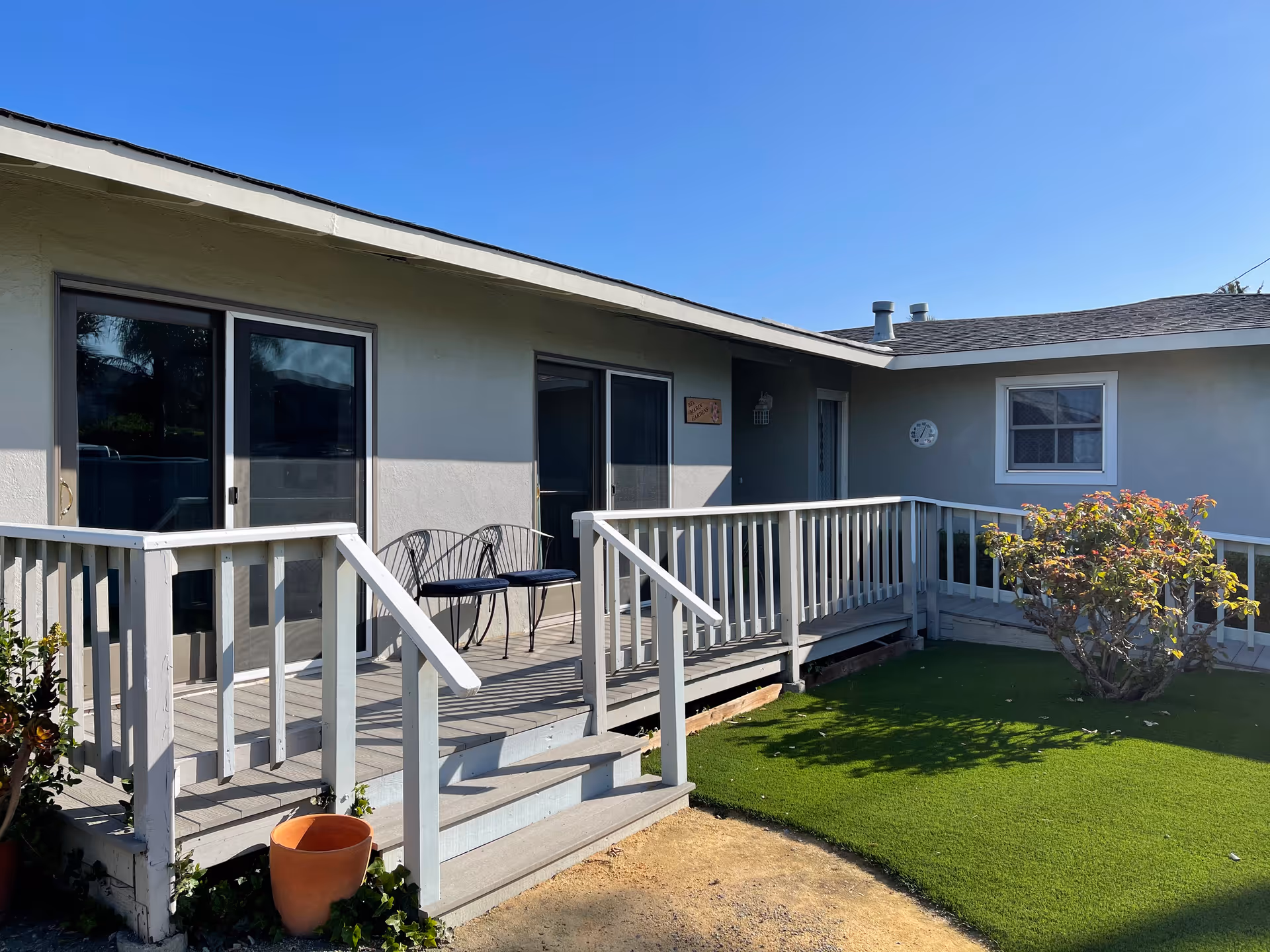 Exterior view of a single-story unit with a wooden ramp/porch, sliding glass doors, outdoor chairs, and a small yard under a clear blue sky.