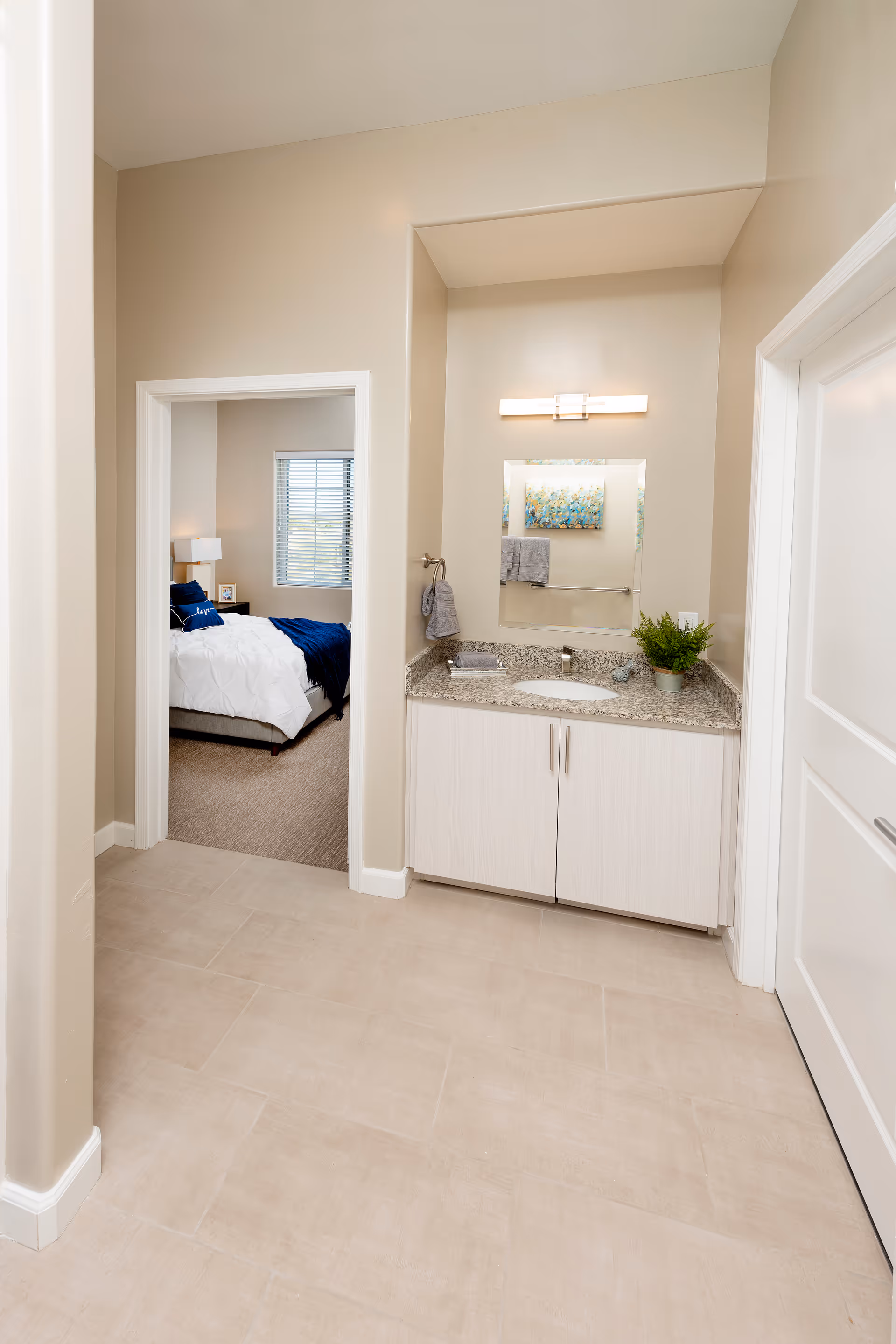 A bright interior space featuring a small vanity area with a granite countertop, a sink, a mirror, and a wall-mounted light fixture. To the left, there is an open doorway leading to a bedroom with a neatly made bed, white bedding, and a blue throw blanket. The bedroom has a window with blinds and a bedside table with a lamp.