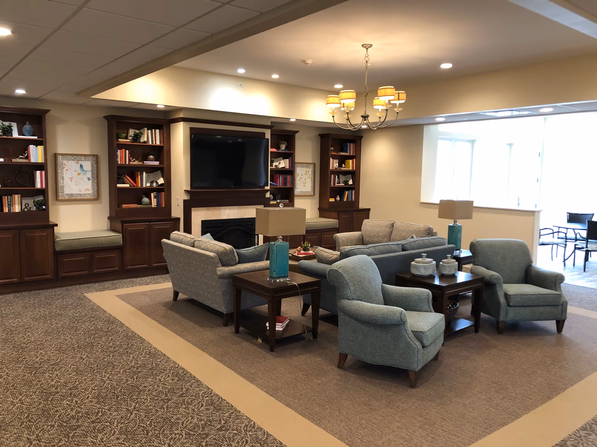 Spacious senior living lounge with sofas and armchairs arranged around tables, built-in bookshelves and a TV over a fireplace.