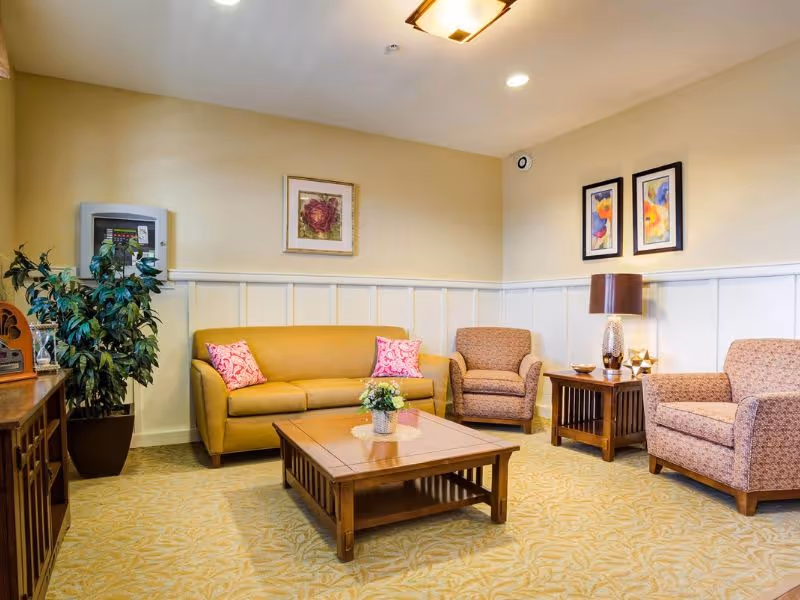 A cozy living room with a mustard yellow sofa adorned with two pink patterned pillows, two beige armchairs, a wooden coffee table with a small flower arrangement, a wooden side table with a lamp and decorative items, framed floral artwork on the walls, a potted plant, and soft yellow patterned carpet.