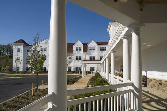 View of the exterior of a senior living facility with white siding and multiple windows, seen from a covered porch with white columns and railing. The porch has rocking chairs and overlooks a landscaped walkway and parking area under a clear blue sky.