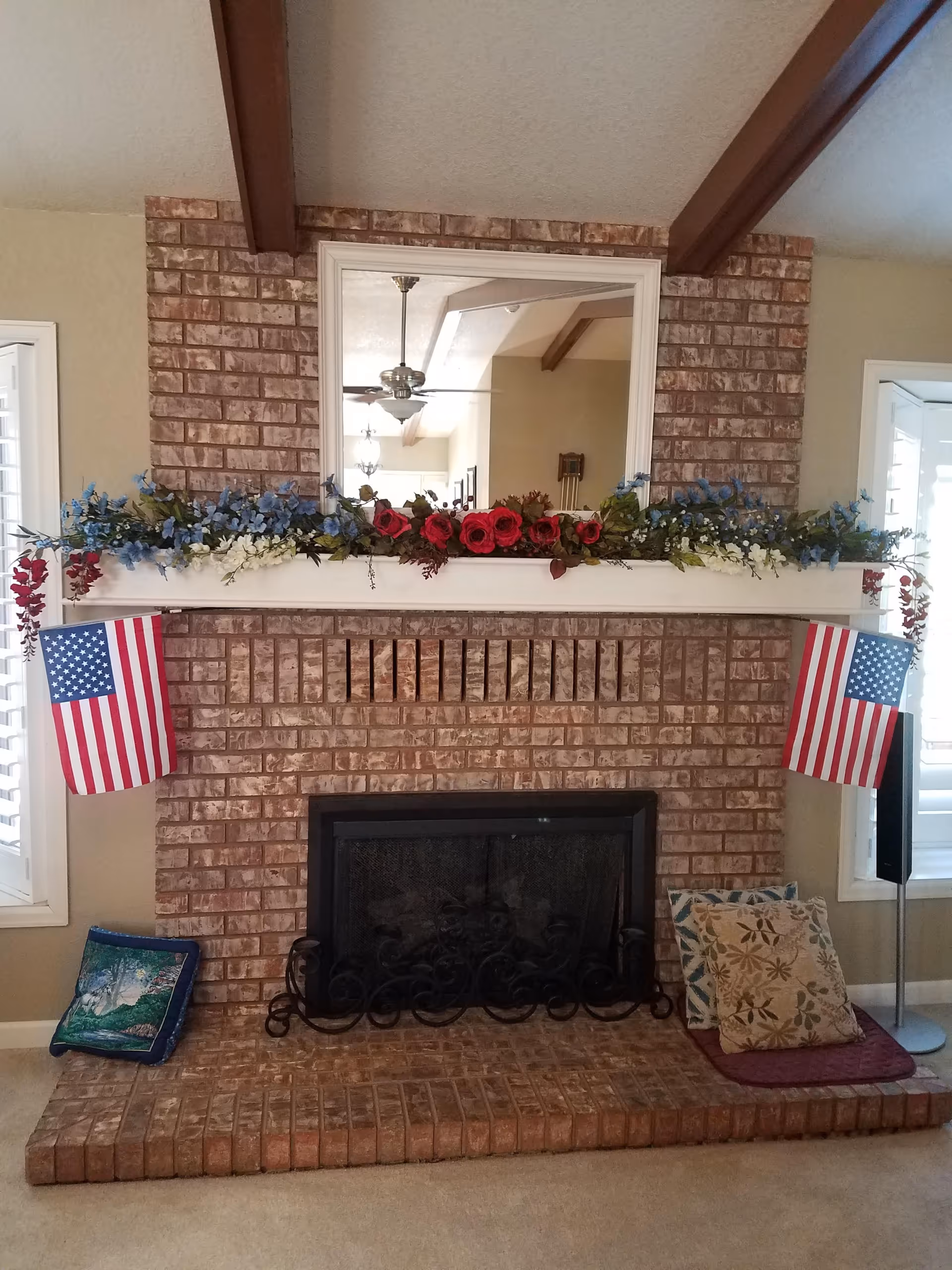 A brick fireplace with a white mantel decorated with red, white, and blue flowers and two small American flags hanging on either side. Above the mantel is a large mirror reflecting a ceiling fan and part of the room. There are decorative pillows placed on the hearth on both sides of the fireplace.