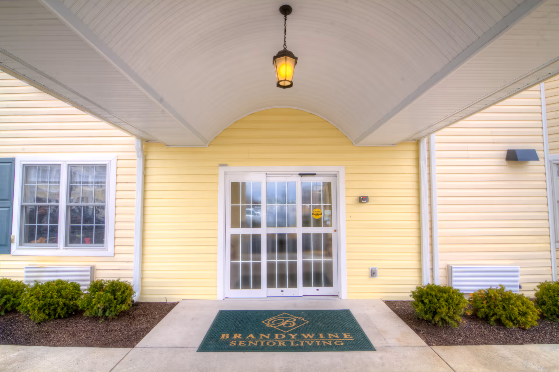 Covered front entrance of a yellow-sided senior living building with glass double doors and a doormat reading Brandywine Senior Living.