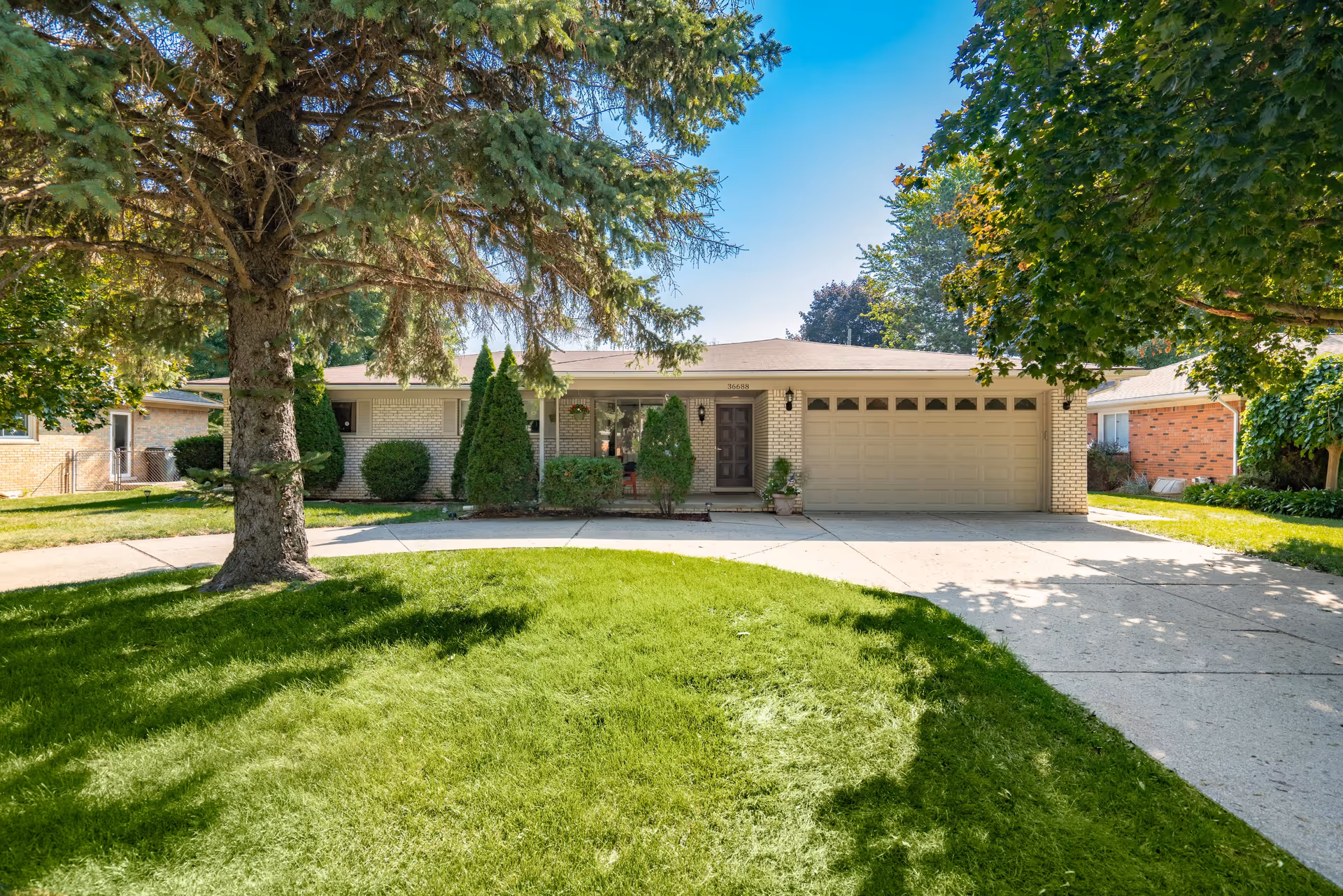 Front exterior view of a single-story brick house with a two-car garage, a driveway, and a well-maintained green lawn with trees providing shade.