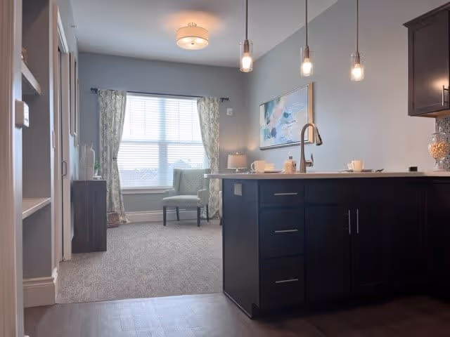 Interior view of a living space in an assisted living facility showing a kitchen island with a sink and dark cabinetry in the foreground. In the background, there is a carpeted area with a green upholstered chair near a window with floral curtains, a small side table with a lamp, and a colorful abstract painting on the wall. Three pendant lights hang above the kitchen island.