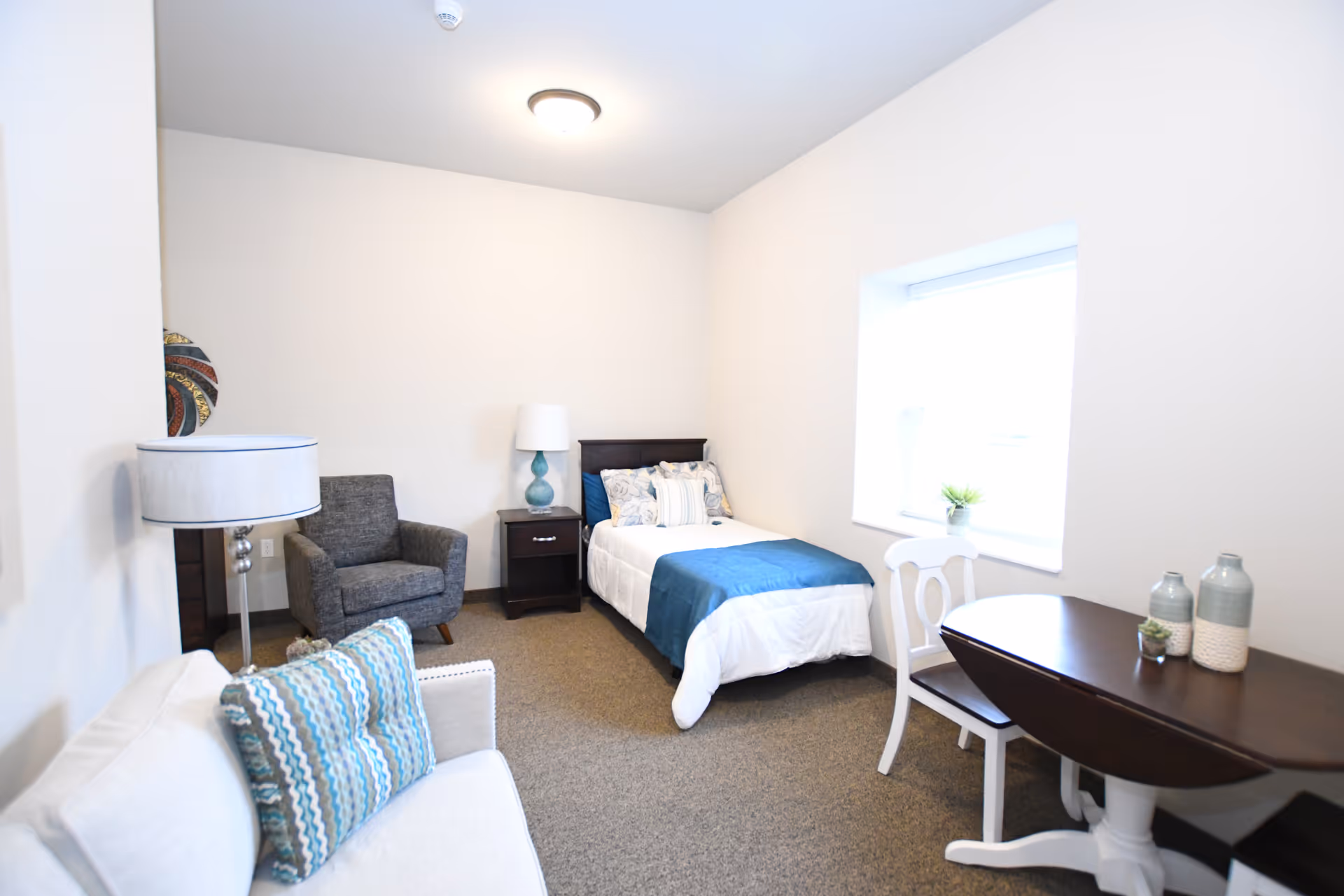 A bright and cozy bedroom with a single bed dressed in white and blue bedding, a dark wooden nightstand with a blue lamp, a gray armchair, a white sofa with a patterned pillow, and a dark wooden table with a white chair near a window with a small plant on the windowsill.