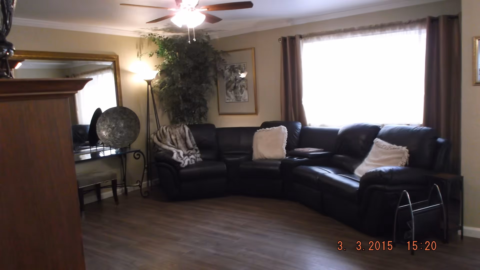 Living room with a black leather sectional sofa adorned with white pillows and a throw blanket. There is a large window with curtains letting in natural light, a tall floor lamp, a large framed mirror on the wall, a decorative plant, and a small side table.