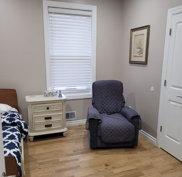 A cozy room with a wooden bed partially visible on the left, a white nightstand with tissue box and small decorative item, a window with white blinds, a comfortable dark gray recliner chair with a quilted cover, and a framed botanical picture on the wall next to a closed white door.