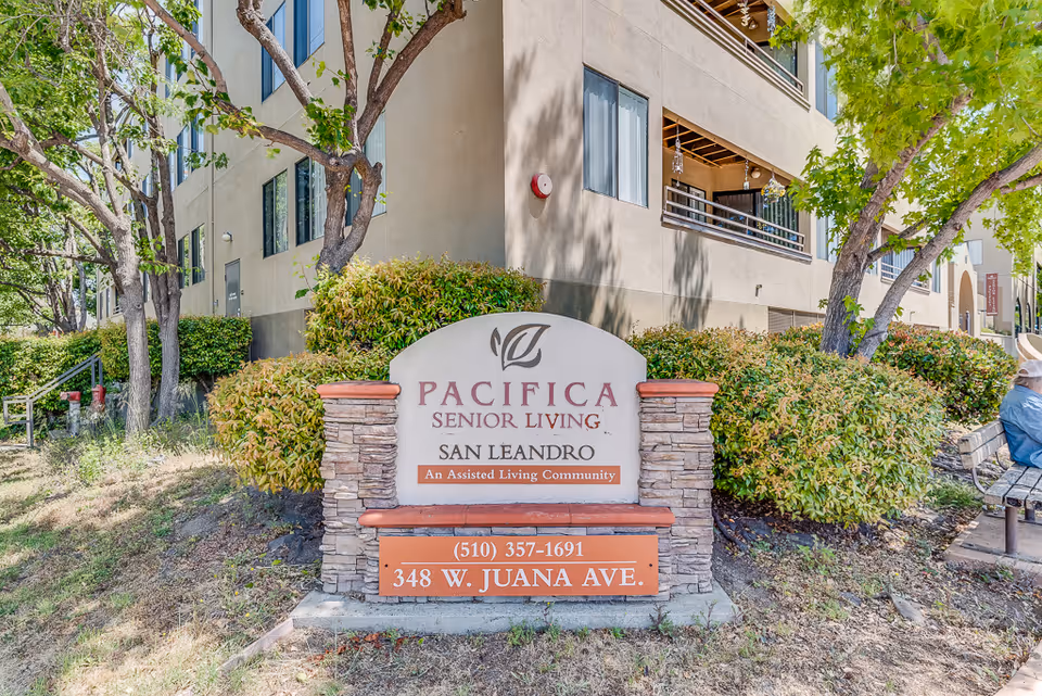 A stone entrance sign reading "Pacifica Senior Living San Leandro" sits in landscaped grounds with the building and trees behind it.