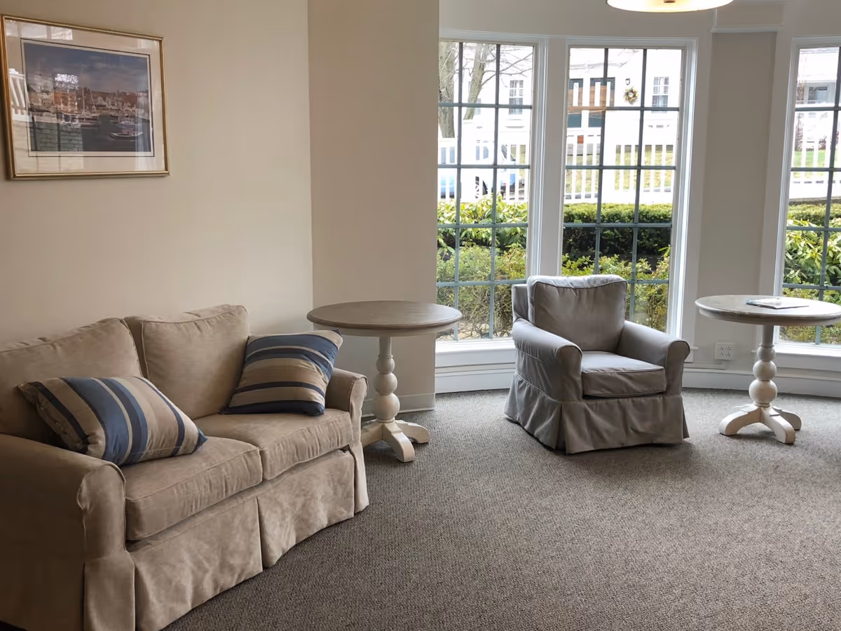 A cozy sitting area with a beige sofa adorned with two striped pillows, a matching armchair, and two round white side tables. Large windows behind the furniture provide a view of greenery and a building outside. A framed picture hangs on the wall above the sofa.