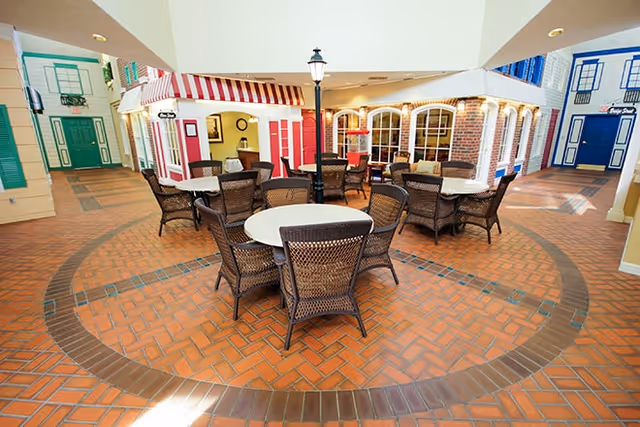 Indoor common area designed to resemble a small village street with brick flooring, round tables with wicker chairs, storefront facades with windows and doors, and a street lamp in the center.