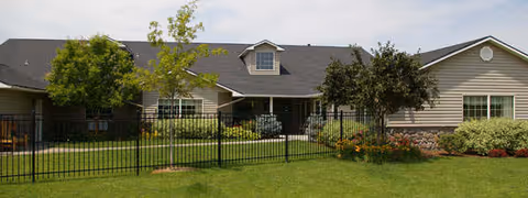 Single-story assisted living building with a fenced front lawn, shrubs, and a covered central entrance.
