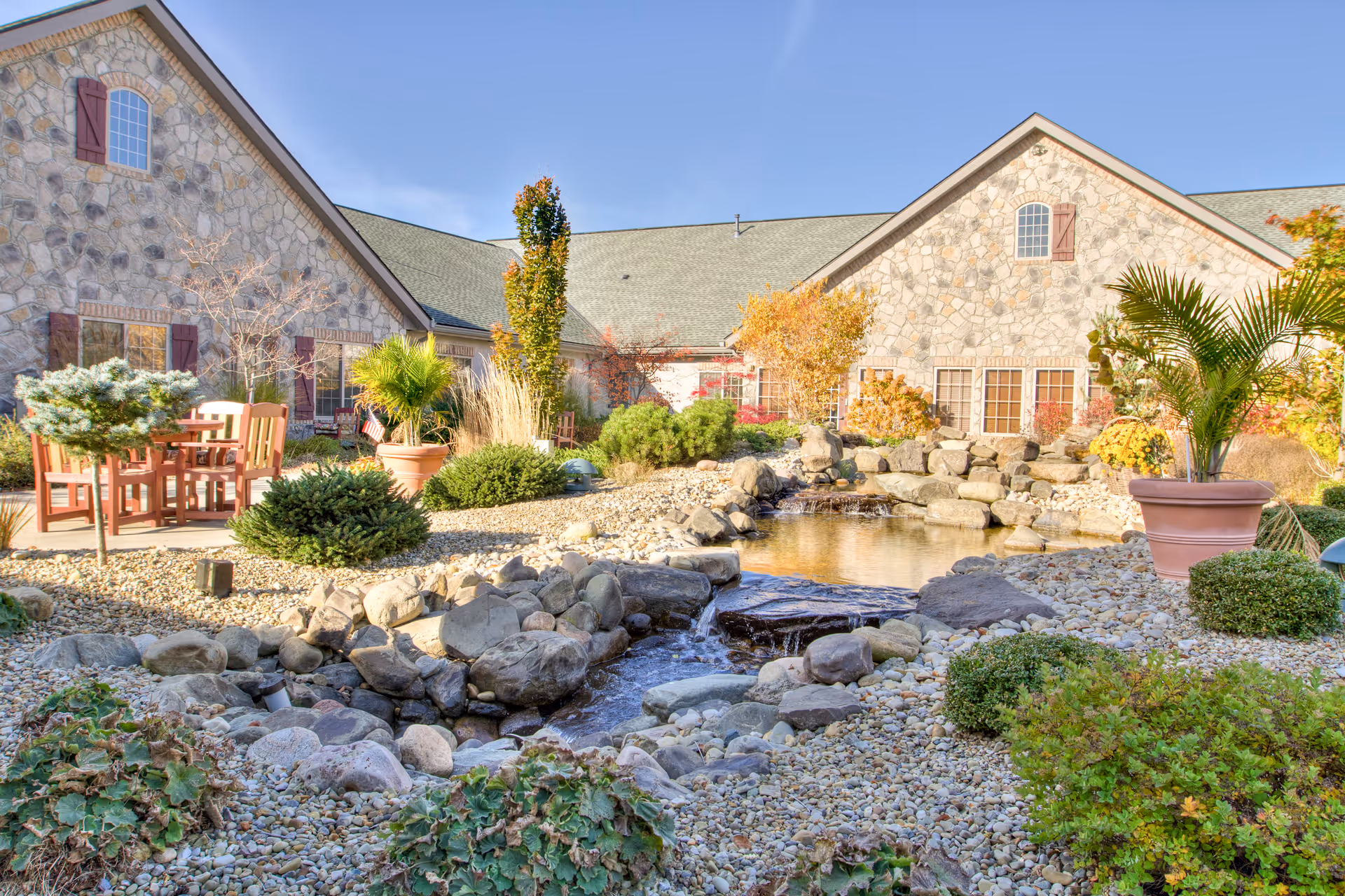 Outdoor garden area at Heritage Senior Living of Marysville featuring a small pond with a waterfall surrounded by rocks, various shrubs, potted plants, and wooden chairs and tables. The background shows a stone building with multiple windows and a clear blue sky.