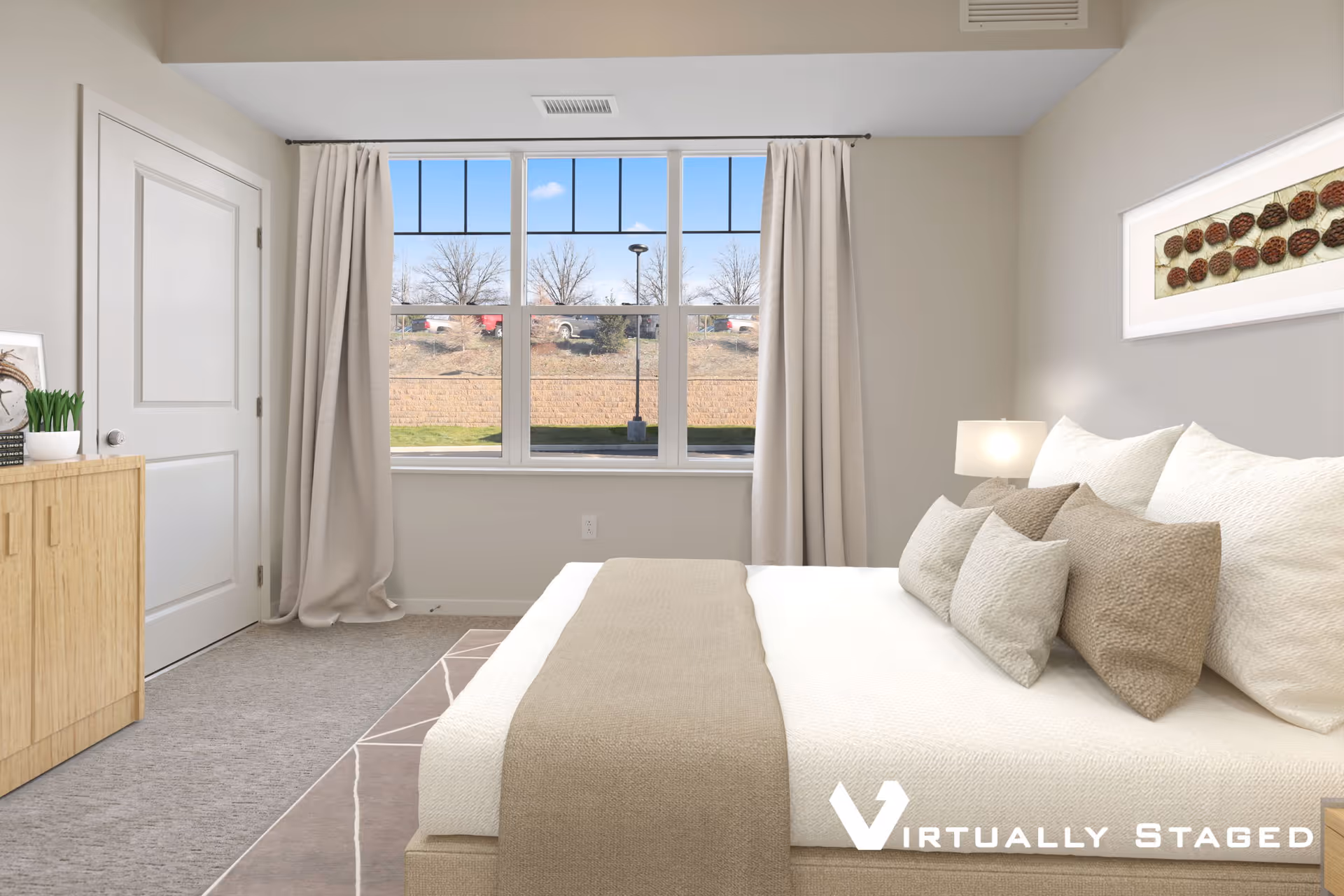 A neatly arranged bedroom with a large window showing an outdoor view. The bed is made with white and beige bedding and multiple pillows. There is a wooden dresser with a small plant and framed picture on the left side. Beige curtains frame the window, and a modern wall art piece hangs above the bed. The room has neutral tones and a carpeted floor.