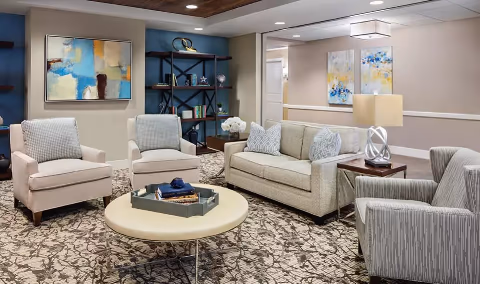 A cozy living room area with a beige sofa, two beige armchairs, and a gray patterned armchair arranged around a round coffee table with books and a decorative tray. The room features a patterned carpet, a wooden bookshelf with decorative items, abstract paintings on the walls, and a table lamp on a side table.