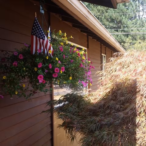 Exterior side view of a brown building with a hanging basket of pink and yellow flowers and an American flag. There is a leafy bush or small tree in the foreground and trees in the background.