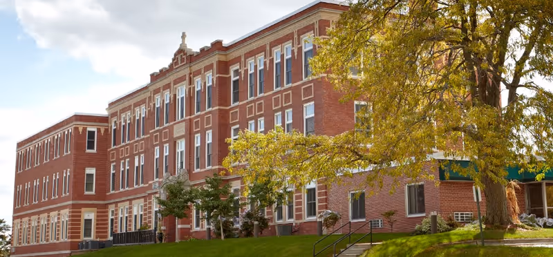 A large, three-story red brick building with white trim and many windows, surrounded by green grass and trees with yellowing leaves under a partly cloudy sky.