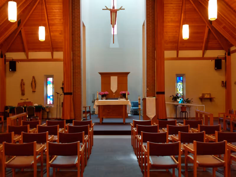 Interior view of a small chapel with rows of wooden chairs facing an altar, a hanging crucifix, stained-glass windows, and warm wood paneling.