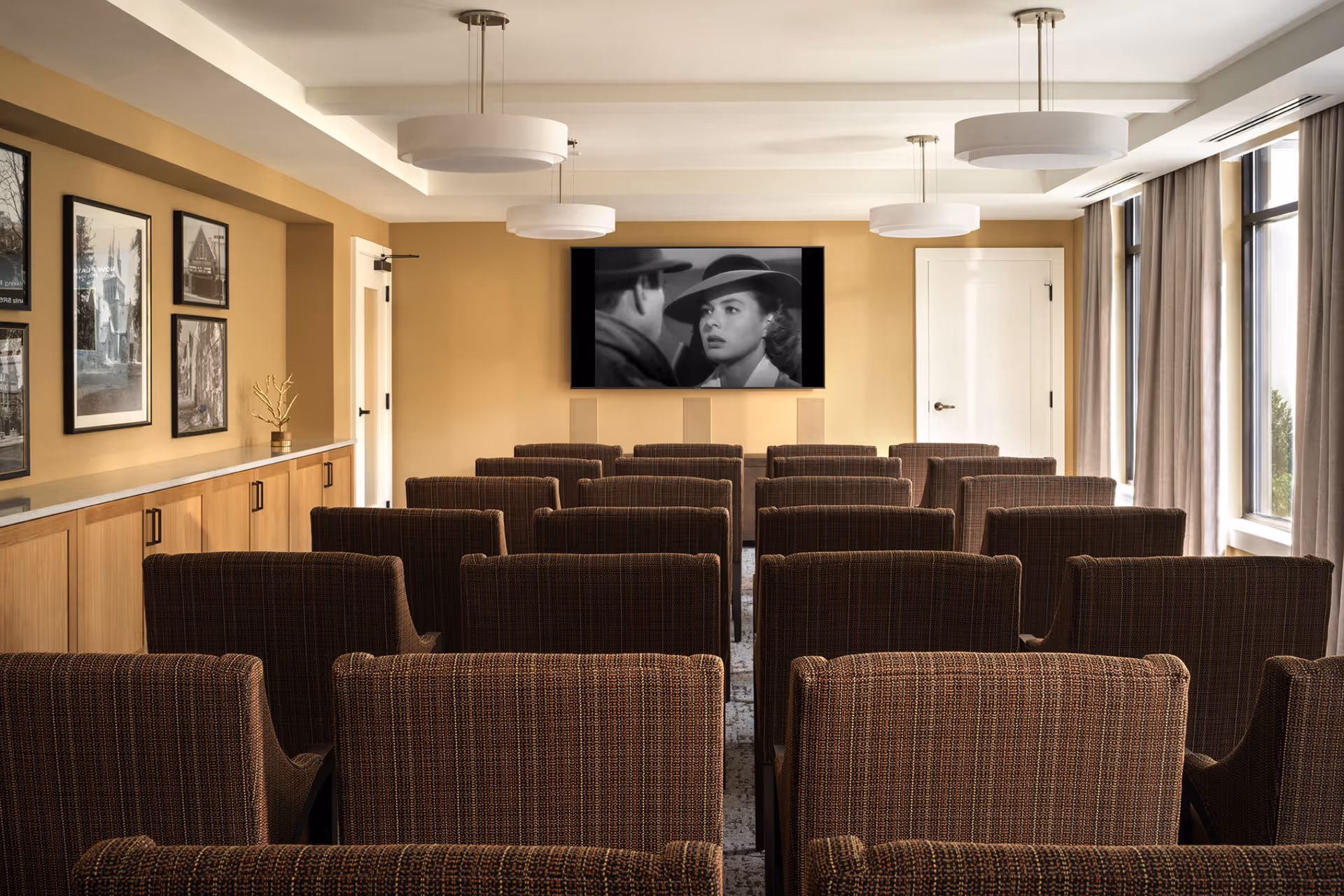 A small theater room with multiple rows of brown upholstered chairs facing a wall-mounted flat screen TV showing a black and white movie scene. The room has beige walls, three modern ceiling lights, framed black and white photographs on the left wall, and large windows with curtains on the right side.