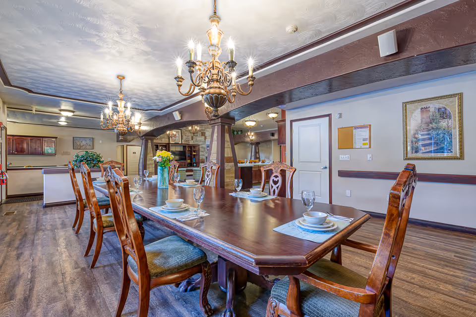 A spacious dining room with a long wooden table set with plates, bowls, glasses, and placemats. The room features ornate wooden chairs, two large chandeliers hanging from a textured ceiling, and a vase with flowers as a centerpiece. The background shows a kitchen area and a framed picture on the wall.