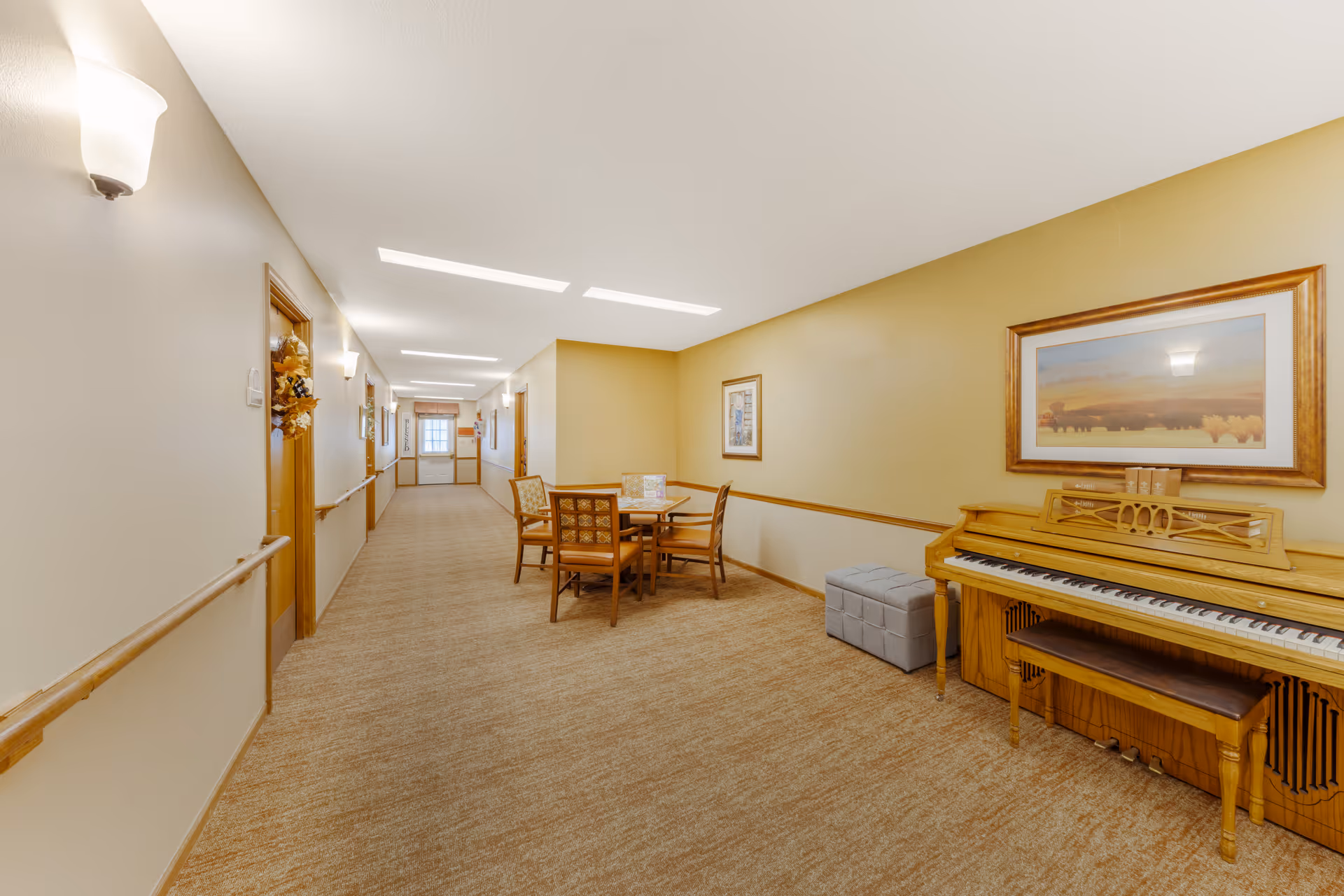 Bright hallway/common area in a senior living facility with a piano, table and chairs and room doors along the corridor.