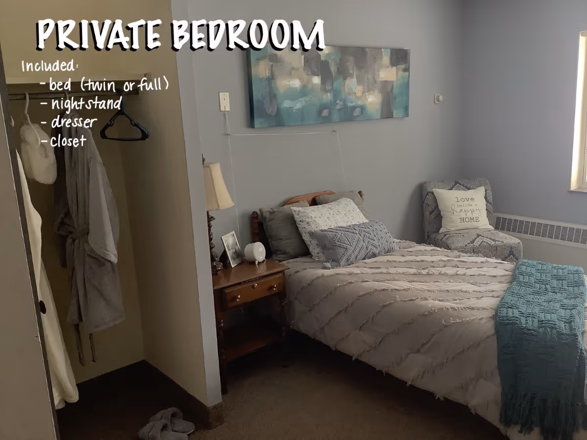 A private bedroom with a bed made up with gray and white bedding and multiple pillows. There is a wooden nightstand with a lamp, a small clock, and a framed photo. To the left, there is an open closet with hanging robes and hangers. A cushioned chair with a pillow that says 'love builds a happy HOME' is placed near a window with a radiator below it. A teal throw blanket is folded at the foot of the bed. A large abstract painting hangs above the bed. Text on the image lists included items: bed (twin or full), nightstand, dresser, closet.