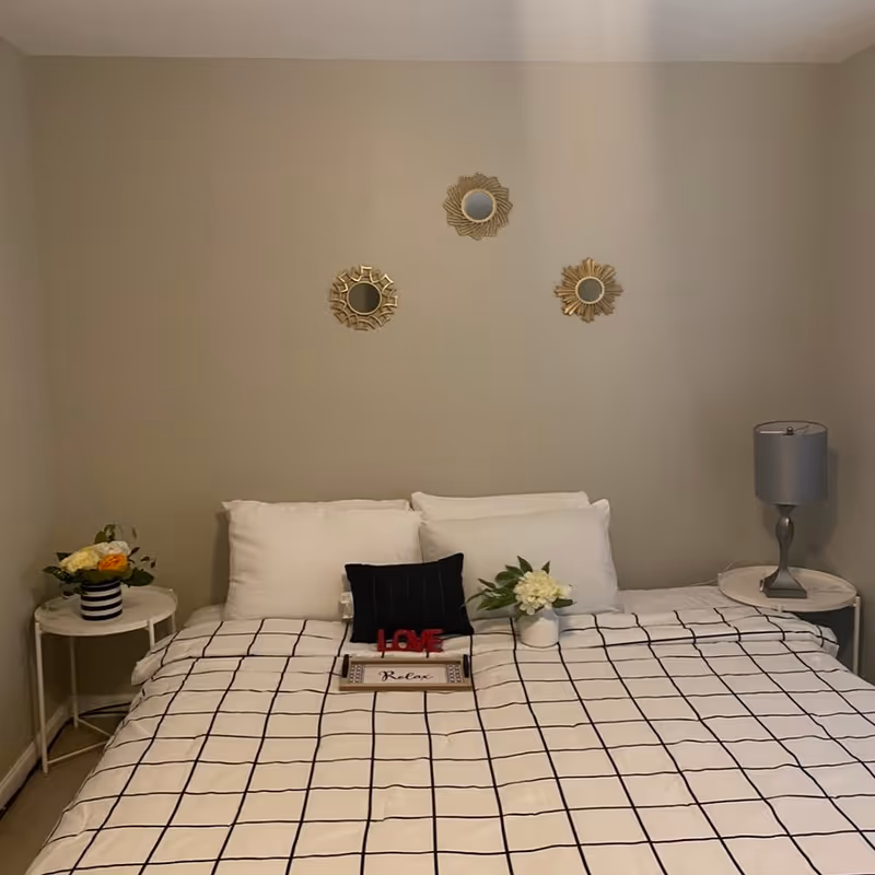Bedroom with a neatly made bed covered by a black-and-white checkered duvet, two pillows, decorative wall mirrors above, and nightstands with flowers and a lamp.
