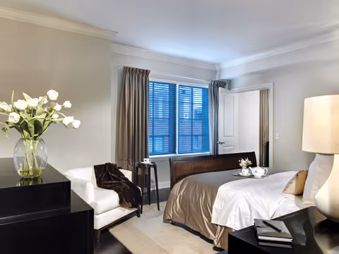 A well-lit bedroom featuring a large bed with white and beige bedding, a bedside table with a lamp, a white armchair with a dark throw blanket, a black dresser with a vase of white flowers, and a window with beige curtains letting in natural light.