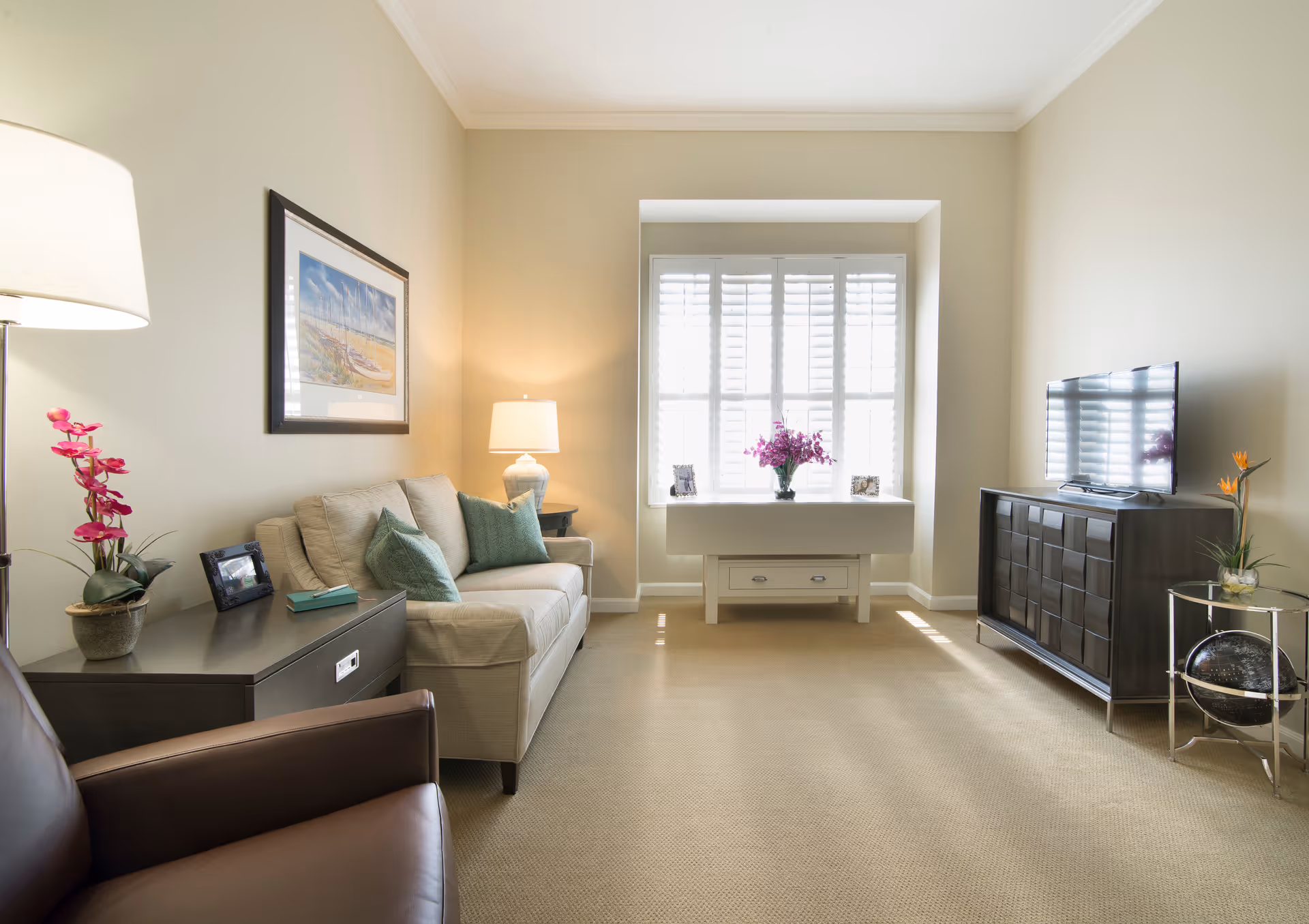 Well-lit living room with a sofa and armchair, TV on a cabinet, a window seat with flowers, and decorative plants and lamps.