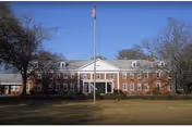 Front exterior view of a large two-story brick building with white trim and multiple windows, an American flag on a flagpole in front, and trees on either side under a clear blue sky.