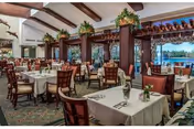 Spacious dining room with multiple tables covered in white tablecloths, set with silverware and small floral centerpieces. The room features large windows letting in natural light, wooden beams on the ceiling, and decorative plants on top of wooden columns.
