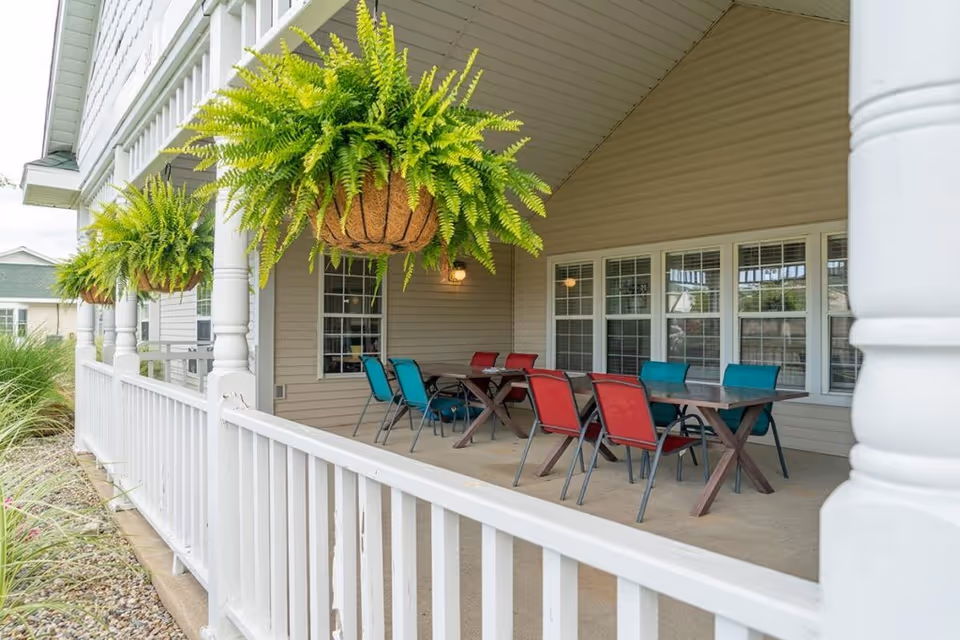 Covered outdoor patio area with hanging green ferns, white railing, and multiple tables surrounded by red and teal chairs, attached to a beige building with several windows.