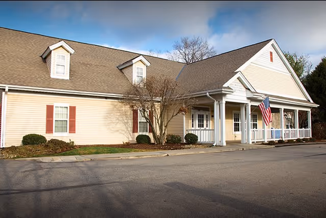 Exterior view of a single-story building with beige siding, white trim, and a pitched roof. The building has several windows with red shutters, a covered porch with white columns, and an American flag hanging near the entrance. There are some bushes and a tree in front of the building, and a paved driveway or parking area in the foreground under a partly cloudy sky.