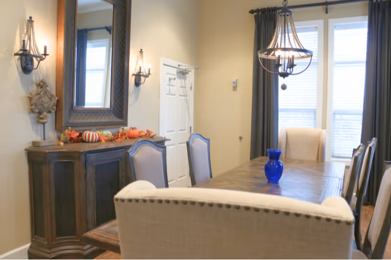 A dining room with a wooden table surrounded by upholstered chairs. A blue vase is placed on the table. The room features a chandelier hanging from the ceiling, two windows with dark curtains, a wooden sideboard with decorative items including small pumpkins and a large mirror above it, and wall sconces on either side of the mirror.