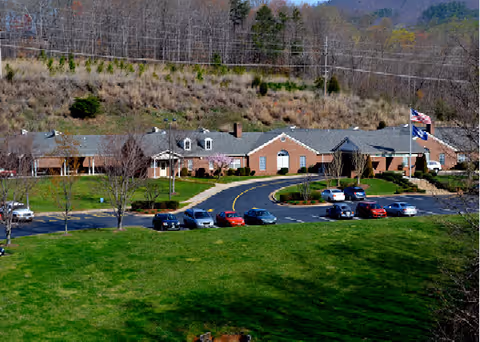 Front view of a single-story brick senior living facility with a circular driveway, parked cars, a flagpole, and a large grassy lawn.
