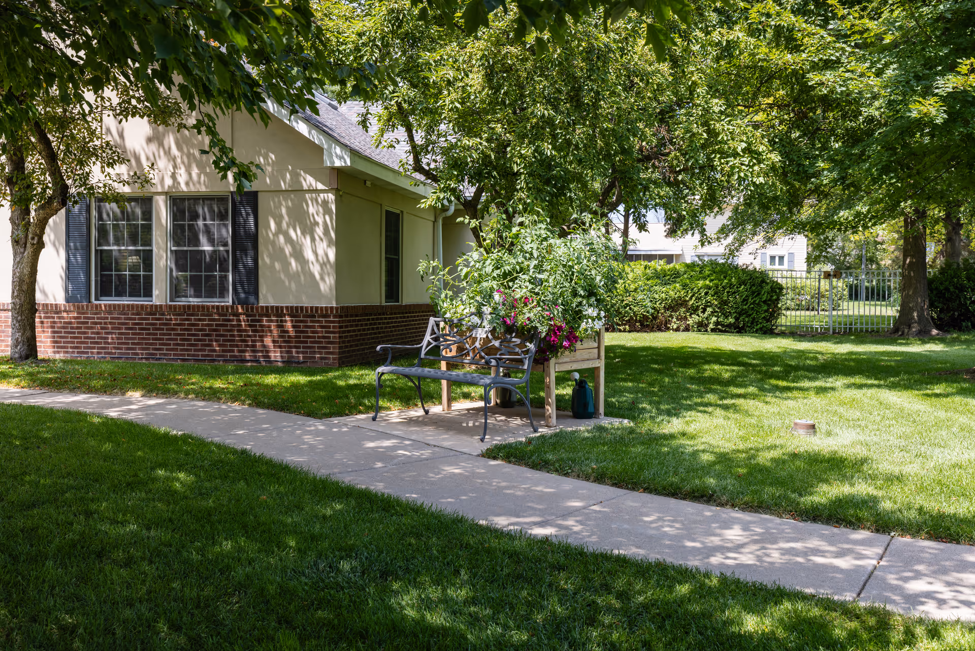 A shaded outdoor area with a concrete pathway, green grass, and trees. There is a metal bench next to a raised garden bed filled with plants and flowers. A building with brick and beige siding and windows is visible in the background.