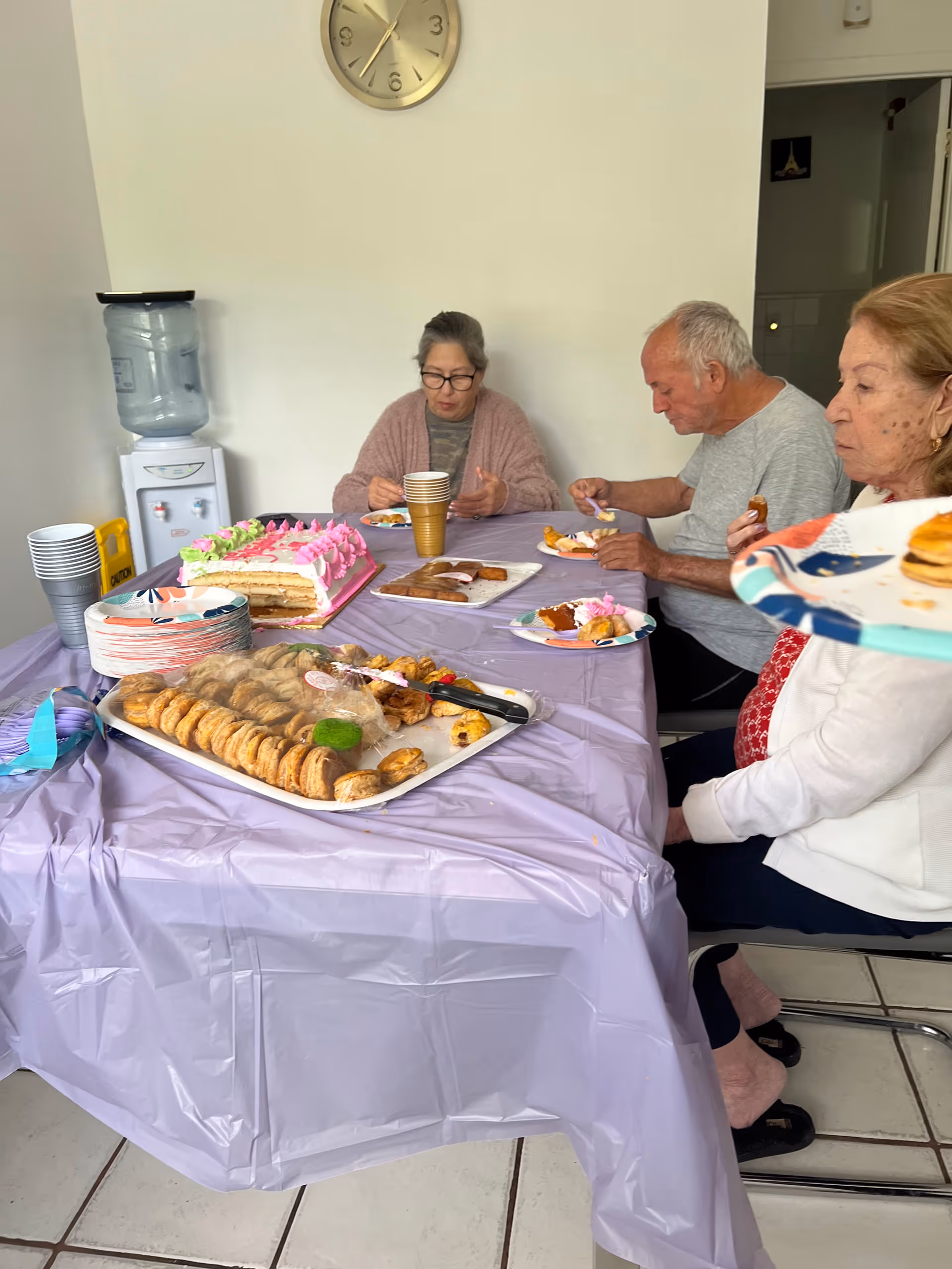 Three elderly people sitting around a table covered with a purple tablecloth, eating cake and pastries. There is a large pink and white cake, plates of cookies, and disposable cups on the table. A water dispenser and a clock on the wall are visible in the background.