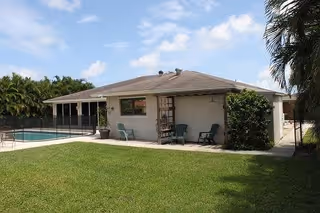 Single-story building with a sloped roof, surrounded by a well-maintained grassy lawn. There are two chairs and a small table on a concrete patio area next to the building. In the background, there is a fenced swimming pool area with palm trees and other greenery around.
