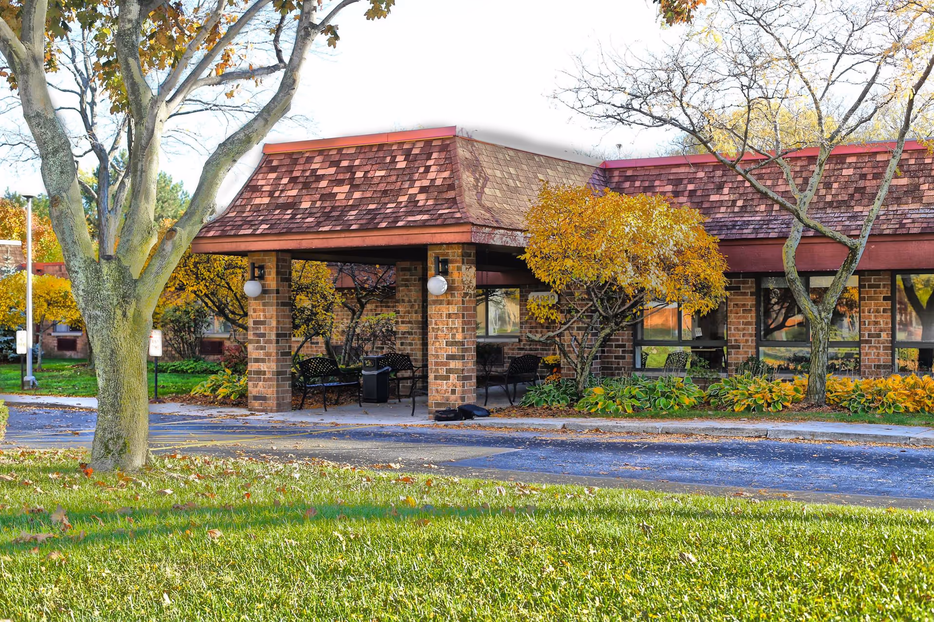 Exterior view of Arbor Inn showing a brick building with a covered entrance supported by brick columns. There are benches under the covered area, trees with autumn foliage, and a grassy lawn in the foreground.
