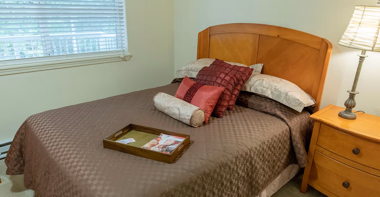 A neatly made bed with a brown quilted bedspread, several decorative pillows in shades of red and beige, and a wooden headboard. A wooden tray with magazines is placed on the bed. To the right of the bed is a wooden nightstand with two drawers and a lamp with a white shade. A window with white blinds is visible on the left side of the image.
