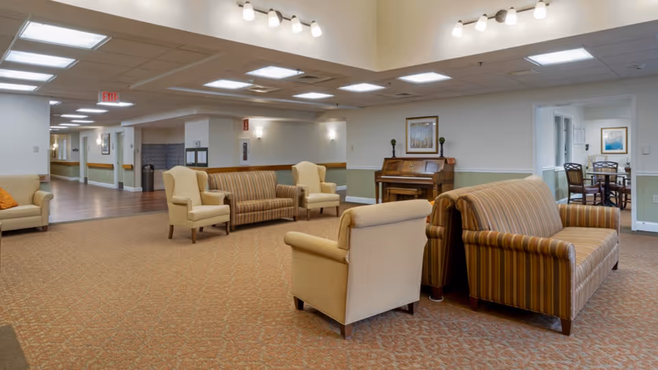 A spacious senior living facility common area with beige and striped upholstered armchairs and sofas arranged on a patterned carpet. There is a wooden piano against the wall with framed artwork above it. The room is well-lit with ceiling lights and has a hallway leading to other rooms.