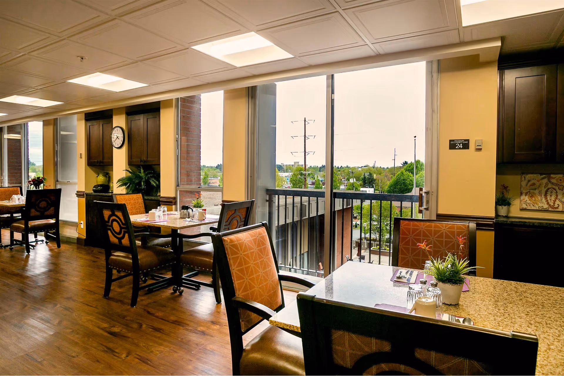 Bright dining room with tables and chairs overlooking an outdoor balcony and treed view through large windows.