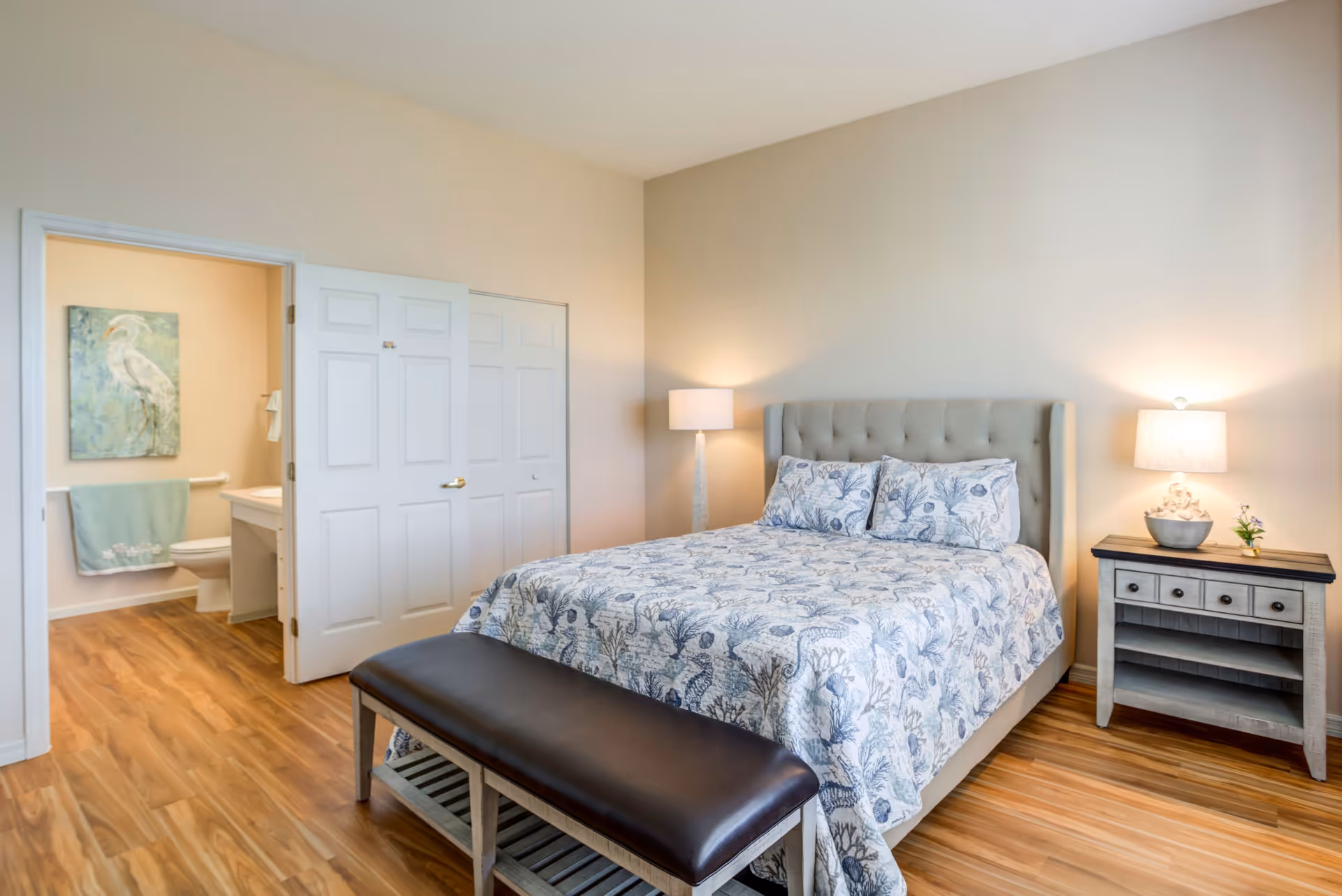 A bedroom with a bed covered in a blue and white patterned quilt, two matching pillows, a padded bench at the foot of the bed, a nightstand with a lamp and small plant, and an open door leading to a bathroom with a toilet, sink, towel, and artwork of a bird on the wall.