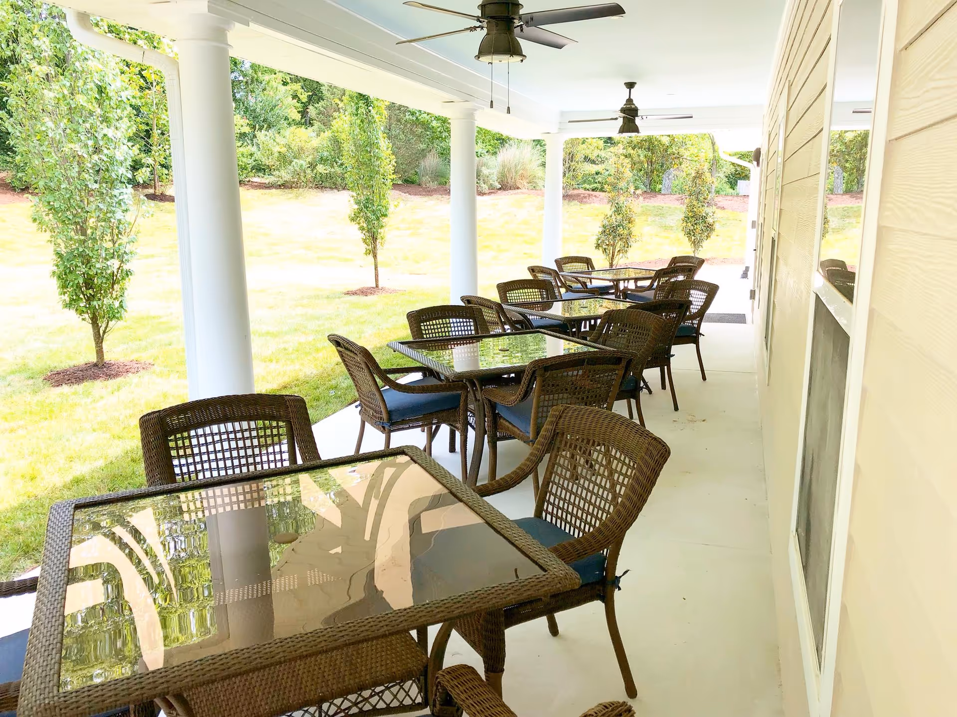 Covered outdoor patio area with multiple glass-top tables and wicker chairs with blue cushions, ceiling fans above, and a grassy area with small trees beyond the patio.